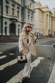 Stylish woman in a coat and sunglasses crossing the street in an urban environment.